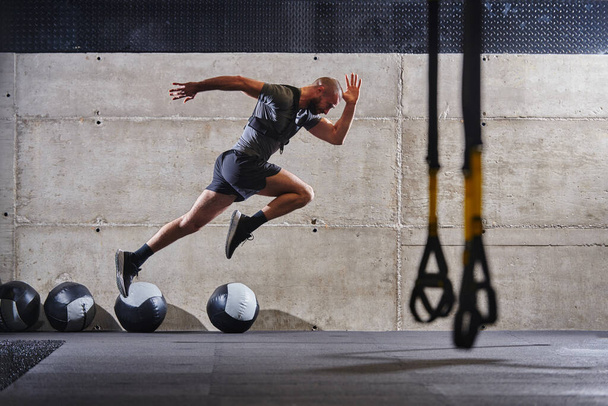 Ein muskulöser Mann, der beim Springen in einem modernen Fitnessstudio in die Luft gejagt wird und seine Athletik, Kraft und Entschlossenheit durch eine intensive Fitness-Routine demonstriert. - Foto, Bild
