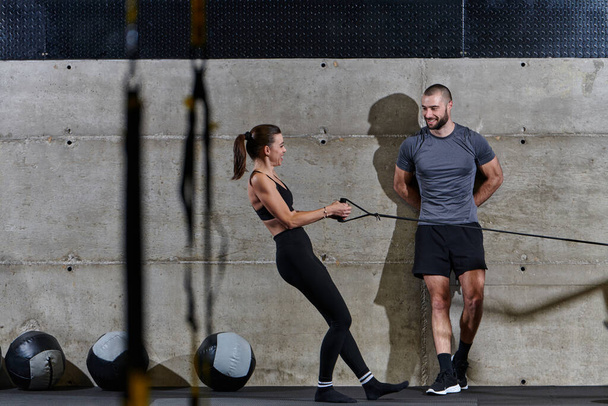 A muscular man assisting a fit woman in a modern gym as they engage in various body exercises and muscle stretches, showcasing their dedication to fitness and benefiting from teamwork and support. - Foto, afbeelding