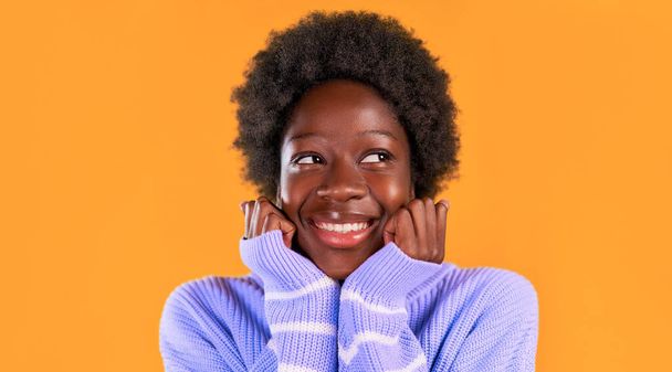 Afričanky americká mladá žena s afro účes stojící v modrém svetru na jasně oranžovém pozadí roztomile dát její pěsti pod bradu. - Fotografie, Obrázek