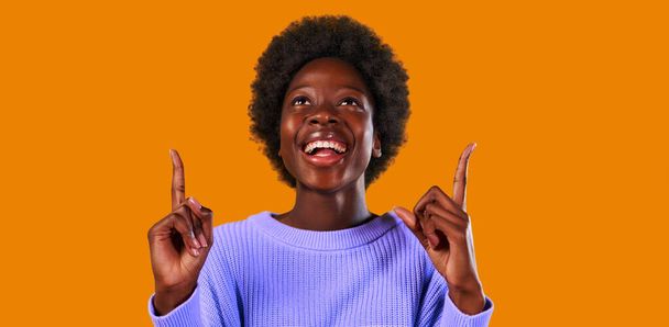Afroamerikanische junge Frau mit Afro-Frisur, die in einem blauen Pullover vor leuchtend orangefarbenem Hintergrund steht, zeigt mit erhobenem Daumen nach oben, um ein Werbeprodukt einzufügen. - Foto, Bild