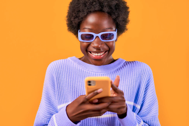 Afroamerykanka młoda kobieta z afro stylizacji włosów stojących w niebieskim swetrze i okulary na jasnopomarańczowym tle przy użyciu telefonu i uśmiechając się radośnie. - Zdjęcie, obraz