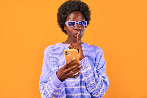 Giovane donna afro-americana con acconciatura afro in piedi in un maglione blu e occhiali su uno sfondo arancione brillante utilizzando il telefono e mostrando un gesto di mettere tranquillamente un dito alla bocca. - Foto, immagini