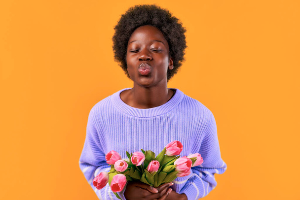 Mujer joven afroamericana con peinado afro de pie en un suéter azul sobre un fondo naranja brillante con un ramo de tulipanes. Día de la Mujer. - Foto, imagen