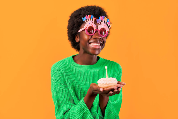 Αφροαμερικανή νεαρή γυναίκα με αφρο χτένισμα σε αστεία γυαλιά με χαρούμενα γενέθλια επιγραφή στέκεται σε ένα πράσινο πουλόβερ σε ένα φωτεινό πορτοκαλί φόντο φυσάει ένα κερί σε ένα cupcake. - Φωτογραφία, εικόνα