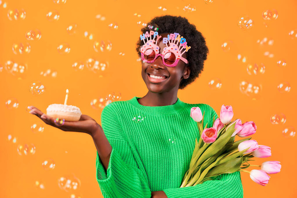 African American young woman with afro hair styling in funny glasses with happy birthday inscription standing on a bright orange background with soap bubbles with a cake and a candle and tulips. - Foto, Imagen