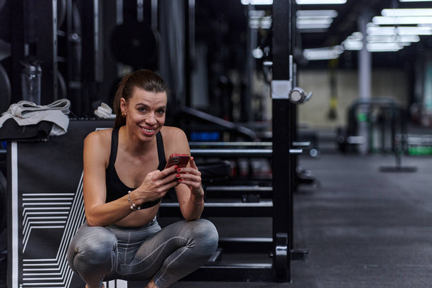A fit woman in the gym taking a break from her training and uses her smartphone, embracing the convenience of technology to stay connected. - Фото, изображение