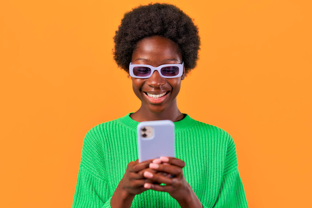 アフリカ系アメリカ人の若いです女性とともにアフロヘアスタイリング立っています緑のセーターで明るいオレンジの背景に眼鏡をかけ、携帯電話を使用して. - 写真・画像