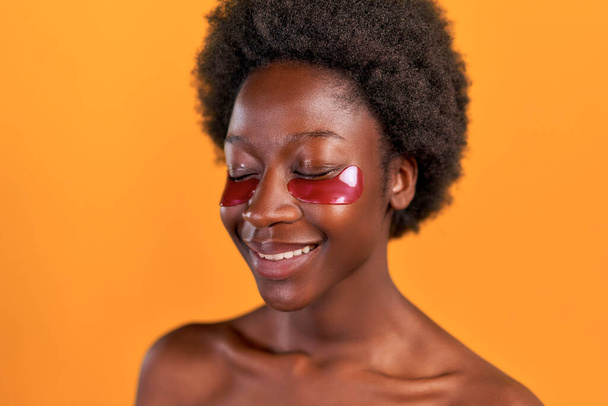 Αφρο-Αμερικανίδα νεαρή γυναίκα με αφρο-μαλλιά κάνει την πρωινή της ρουτίνα χρησιμοποιώντας ενυδατικά μπαλώματα ματιών που απομονώνονται σε πορτοκαλί φόντο. Περιποίηση δέρματος και θεραπείες spa. - Φωτογραφία, εικόνα