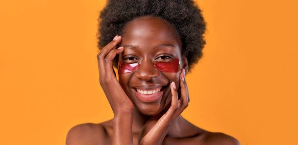 African American young woman with afro hair doing her morning routine using moisturizing eye patches isolated on orange background. Skin care and spa treatments. - Fotó, kép