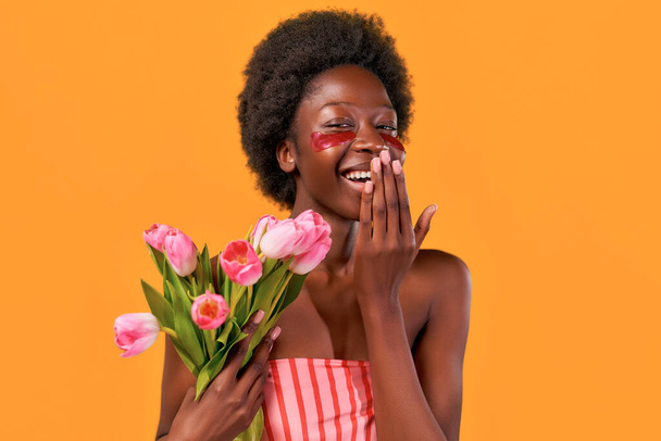 Afroamerikanische junge Frau mit Afro-Haaren mit feuchtigkeitsspendenden Augenklappen, die einen Strauß Tulpen auf orangefarbenem Hintergrund halten. Hautpflege und Wellnessanwendungen. - Foto, Bild