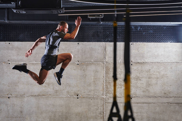Ένας μυώδης άνδρας που αιχμαλωτίζεται στον αέρα καθώς πηδά σε ένα σύγχρονο γυμναστήριο, επιδεικνύοντας τον αθλητισμό, τη δύναμη και την αποφασιστικότητά του μέσα από μια ρουτίνα γυμναστικής υψηλής έντασης. - Φωτογραφία, εικόνα