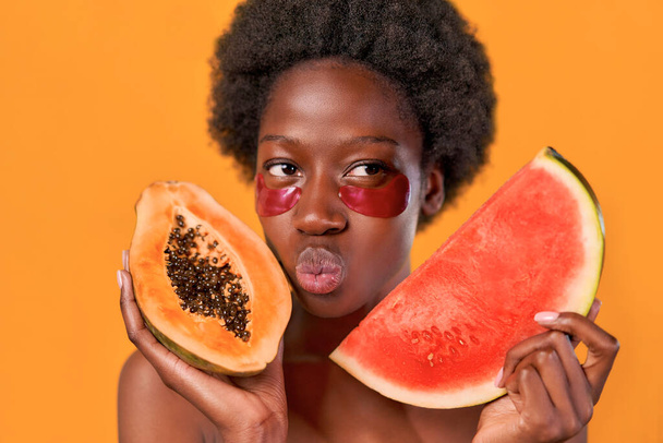 Mujer joven afroamericana con cabello afro usando parches humectantes para los ojos, sosteniendo papaya y sandía en manos aisladas sobre fondo naranja. Tratamientos de cuidado de la piel y spa. - Foto, imagen