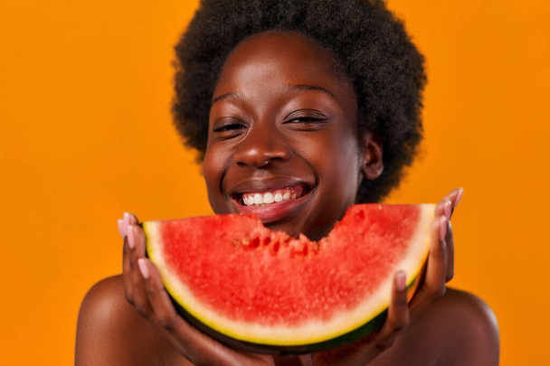 Afro-Amerikaanse jonge vrouw met afrohaar neemt een smakelijke hap sappige watermeloen geïsoleerd op oranje achtergrond. Huidverzorging. Gezonde levensstijl. - Foto, afbeelding