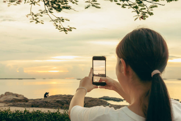 Aasian naiset ottavat kuvia ja videopuheluita merellä ja rannalla aamulla auringonnousun aikaan matkapuhelimillaan, älypuhelimillaan internetiin lähettämistä varten lomalla.. - Valokuva, kuva