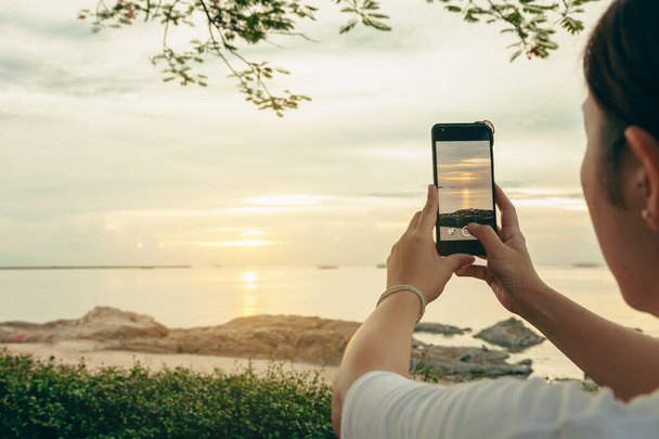 アジアの女性は、休暇中にインターネットに投稿するためのスマートフォンを携帯電話で日の出に海とビーチで写真やビデオ通話を取ります. - 写真・画像
