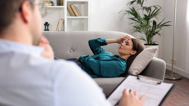 A young woman is lying on a couch at a psychologists appointment. A brunette male psychologist in glasses conducts a psychological session sitting on a chair in his office - Φωτογραφία, εικόνα