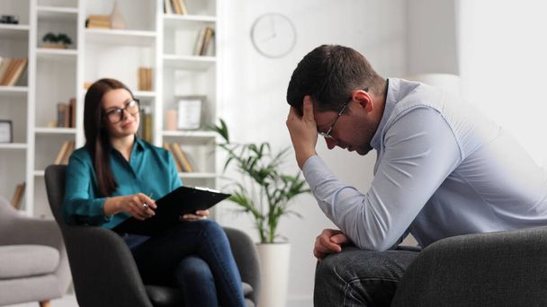 A professional psychologist consults a male patient in a state of stress and depression, who is sitting in a chair in the office. She writes on paper. The concept of a consultant psychologist - Photo, Image