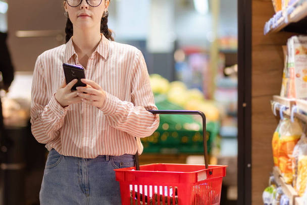Young Caucasian woman holds shopping basket and uses smartphone. Close up. Shopping and consumerism. - Foto, immagini
