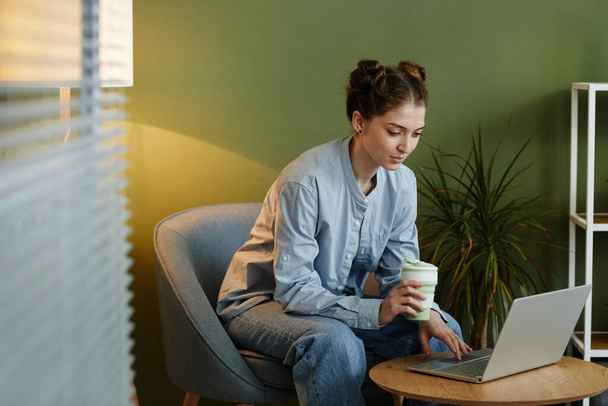 Retrato de mujer joven en ropa casual usando portátil en el rincón del lugar de trabajo contra la pared verde - Foto, Imagen