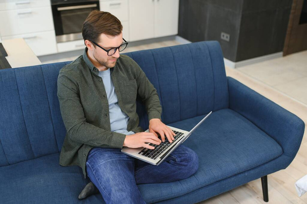 Men working on laptop computer in his room. Home work or study, freelance concept. Young man sitting relaxed on sofa with laptop. Modern businessman using laptop - Photo, Image