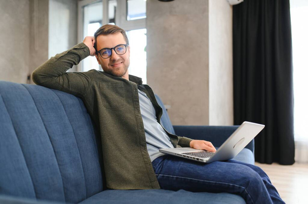 Smiling young man wearing glasses using laptop, sitting in cozy armchair, happy male looking at computer screen, chatting in social network or shopping online, playing game, working at home - Foto, Imagen