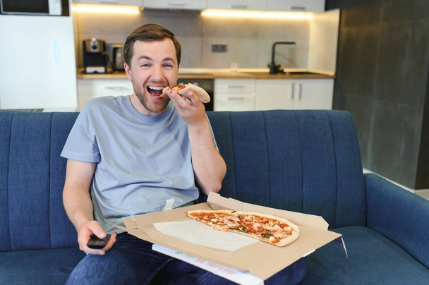 Белый мужчина отдыхает на диване, ест пиццу и смотрит телевизор. Концепция современного успешного образа жизни человека и отдыха после работы. Уютная однокомнатная квартира - Фото, изображение
