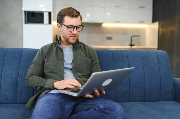 Satisfied handsome young caucasian man in casual stylish clothes, sitting on the couch at home in cozy living room, using a laptop, working or study online, dreaming, smiling. - Fotoğraf, Görsel