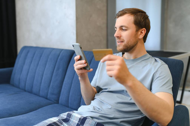 A happy smiling caucasian guy, in casual stylish clothes, sits on the couch at home, holds a phone and a banking card in his hands, makes online payment for purchases, enters a card number. - Фото, зображення