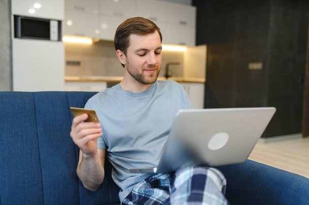 Happy Caucasian man buying things online, using smartphone, laptop and credit card, enjoying shopping in internet. Positive young guy purchasing goods on web, making remote payment. - Photo, image