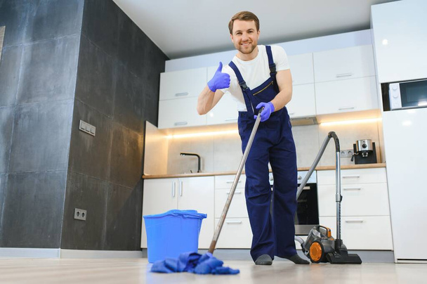 Professional cleaner in blue uniform washing floor and wiping dust from the furniture in the living room of the apartment. Cleaning service concept. - Foto, afbeelding