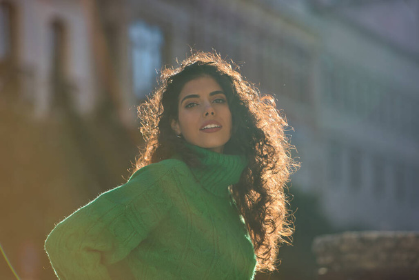 Νέα, όμορφη, μελαχρινή γυναίκα με σγουρά μαλλιά και πράσινο ζιβάγκο πουλόβερ, τυλιγμένο στις ακτίνες του απογευματινού ήλιου. Concept ομορφιά, μόδα, φθινόπωρο, χειμώνας, κρύο, ήλιος. - Φωτογραφία, εικόνα