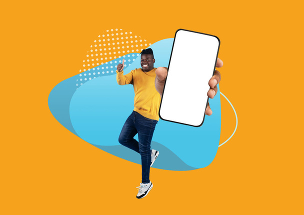 Promoción en línea. Joyful Black Man saltando con un gran teléfono inteligente en blanco en la mano y mostrando Pulgar hacia arriba, alegre afroamericano macho mostrando teléfono móvil con pantalla blanca sobre fondo abstracto, burla - Foto, imagen