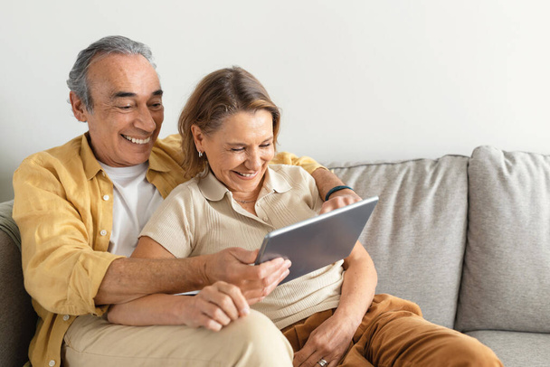 Pensioen en internet leuk concept. Opgewonden oudere echtgenoten met behulp van digitale tablet computer, websurfen of film kijken online, rusten op de bank thuis, vrije ruimte - Foto, afbeelding