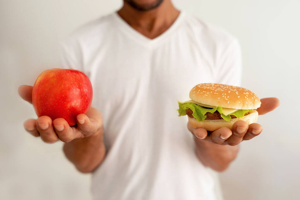 Health Or Junk Food. Cropped Shot Of Black Man Holding Burger And Apple Fruit Choosing Between Healthy Vs Unhealthy Food Against White Wall Background. Cheat Meal And Diet Concept - Foto, imagen