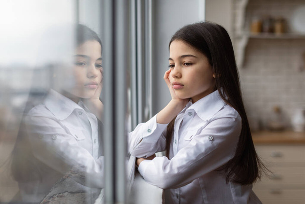 Παιδική κατάθλιψη. Δυστυχισμένη έφηβη που κοιτάζει από το παράθυρο και υποφέρει από δυστυχία και μοναξιά στο σπίτι. Μια μαθήτρια που ποζάρει μόνη της μέσα. Έννοια θέματα ψυχικής υγείας παιδιών - Φωτογραφία, εικόνα