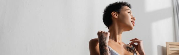young and sexy tattooed woman with short brunette hair touching straps of silk bra while looking away and dreaming near white wall in light bedroom at home, banner - Photo, Image