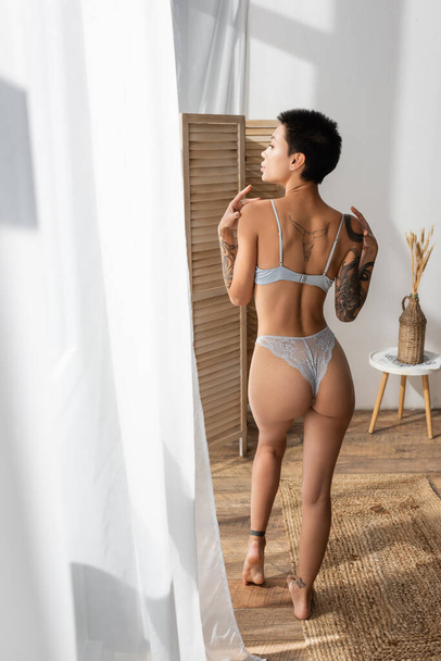 back view of sultry woman with sexy tattooed body wearing lace panties and bra, standing on wicker rug, looking away near white curtain, room divider and vase with spikelets on bedside table  - Foto, Bild