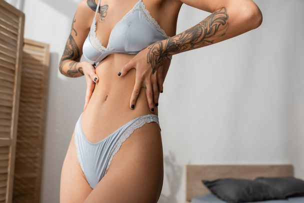 partial view of provocative young woman in grey silk lingerie such as bra and panties touching sexy and slender body with tattooed arms near blurred bed and room divider in bedroom - 写真・画像