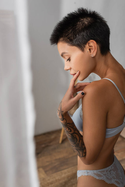 side view of young captivating woman with short brunette hair and tattooed arm touching face while standing in lace panties and bra near white curtain on blurred foreground in bedroom - Foto, immagini