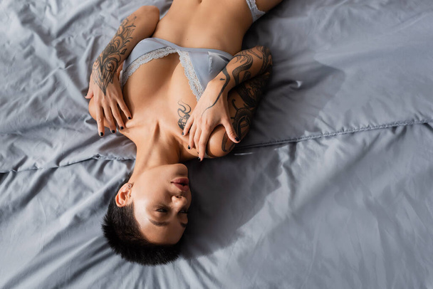 top view of young and provocative woman with short brunette hair and sexy tattooed body laying on grey bedding in silk lingerie in modern bedroom at home - Фото, зображення