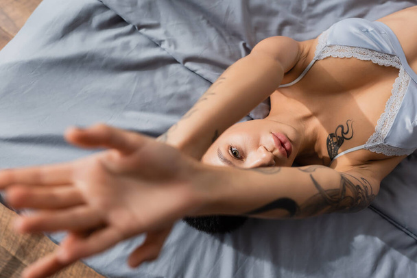 top view of charming and provocative young woman with sexy tattooed body laying in bra with raised hands and looking at camera on grey bedding on blurred foreground - Photo, Image