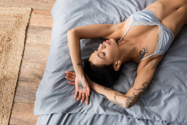 vista superior de la sensual mujer joven en sujetador de seda, con pelo corto morena y cuerpo tatuado sexy tendido en ropa de cama gris cerca de alfombra de mimbre en el suelo en el dormitorio en casa - Foto, Imagen