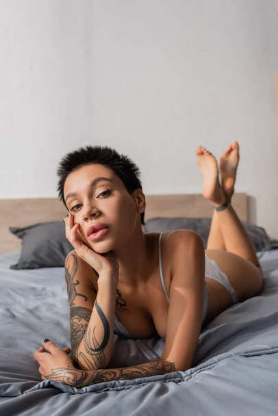 full length of mesmerizing woman with short brunette hair and sexy tattooed body holding hand near face and looking at camera on grey bedding with pillows on blurred background - Photo, Image