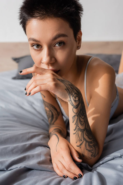 young, charming and provocative woman in lingerie, with short brunette hair and sexy tattooed body holding hand near face and looking at camera on grey bedding at home - Foto, Bild