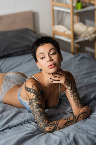 mesmerizing tattooed woman in lingerie, with sexy body and short brunette hair, touching chin and looking away near rack on blurred background in modern bedroom, boudoir photography - Φωτογραφία, εικόνα