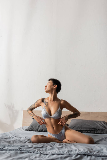 irresistible woman in silk lingerie, with short brunette hair and sexy tattooed body looking away while posing on grey bedding near pillows in modern bedroom, boudoir photography - Foto, afbeelding