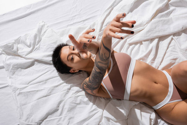 vista superior de mujer tatuada intrigante en lencería beige, con cuerpo sexy y pelo corto morena tendida sobre ropa de cama blanca, señalando con los dedos y mirando a la cámara en el estudio, fotografía erótica - Foto, Imagen