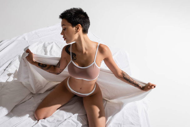 visão de alto ângulo da mulher jovem e sedutora em lingerie bege, com cabelo curto morena e corpo tatuado sexy posando em roupa de cama branca e fundo cinza, arte da sedução - Foto, Imagem