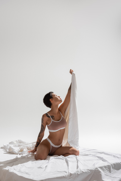 mujer delgada e impresionante en lencería beige, con pelo corto morena y cuerpo tatuado sexy sosteniendo sábana en la mano levantada mientras está sentado sobre ropa de cama blanca y fondo gris, arte de la seducción - Foto, Imagen