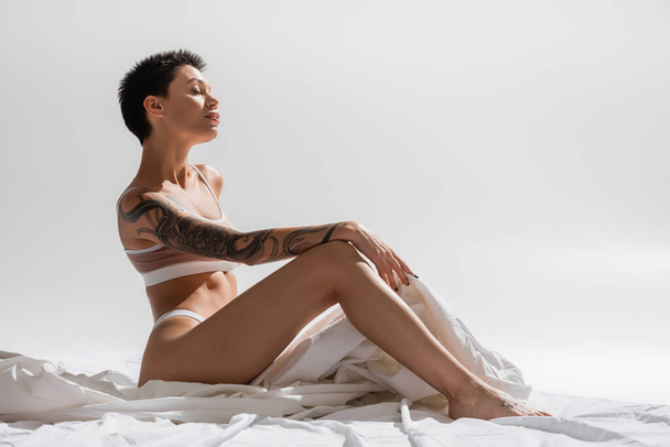 side view of young graceful woman with sexy tattooed body and short brunette hair sitting in beige lingerie with closed eyes on white bedding and grey background in studio, erotic photography - Photo, Image